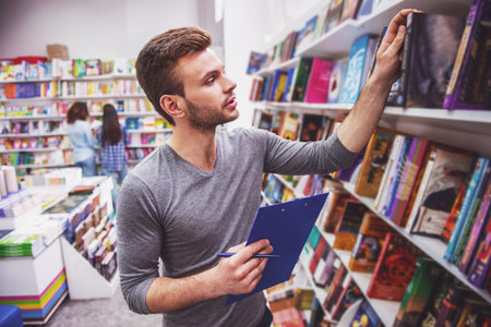 Handsome young man is holding a folder and making notes while choosing a book at the bookshopの写真素材