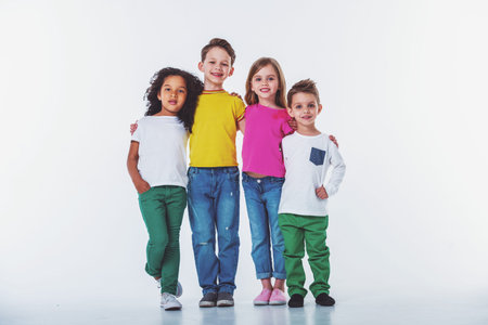 Cute little kids in casual clothes are looking at camera and smiling while standing with their arms...
