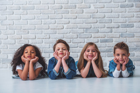 Cute little kids in stylish jeans clothes are leaning on their hands, looking at camera and smiling, lying on the floor against white brick wallの写真素材