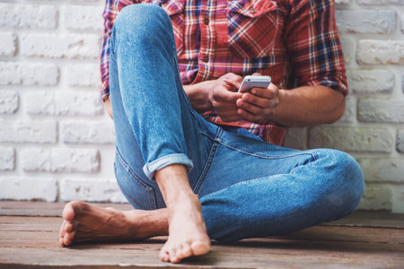 Cropped image of handsome Afro American man in casual clothes using a smartphone while sitting on the floor against white brick wallの写真素材