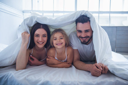 Portrait of beautiful young mother, father and their daughter looking at camera and smiling while lying on bed covered with blanketの写真素材