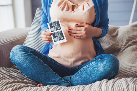 Cropped image of beautiful pregnant woman holding sonogram and keeping one hand on a belly while sitting on couch at homeの写真素材