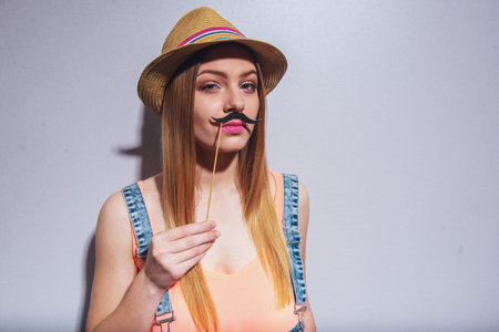 Attractive girl in summer clothes is posing with paper moustache on wood stick and looking at...