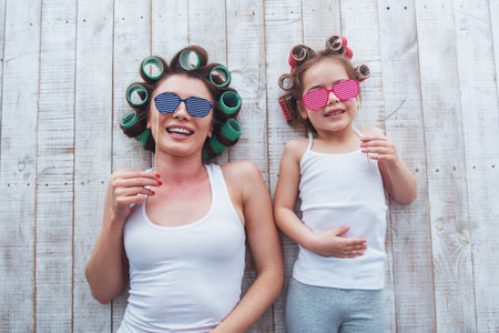 Top view of young mother and her daughter with hair curlers on their heads holding paper glasses on stick and smiling, lying on the floor at homeの写真素材