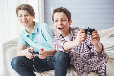 Two attractive teenage boys are playing game console and smiling while sitting on the couch at home