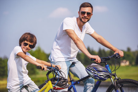 Handsome young dad and his cute little son in casual clothes and sun glasses are looking at camera and smiling while riding bikes in parkの写真素材