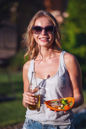 Portrait of beautiful girl in casual clothes and sun glasses holding bottle of beverage and food, looking at camera and smiling while resting outdoorsの写真素材