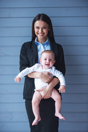 Portrait of beautiful business lady in classic suit and her sweet little baby looking at camera and...