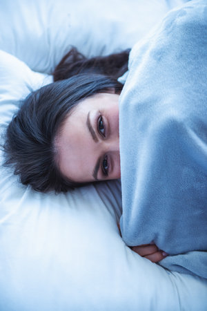 Portrait of beautiful young woman looking at camera and hiding behind blanket while lying in her bed at homeの写真素材