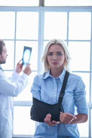 Beautiful female patient with injured arm is holding her bandage and looking at camera, in the background doctor is examining X-ray pictureの写真素材