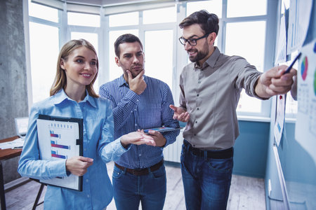 Beautiful business people are studying documents, talking and smiling while working in office. Handsome man is pointing to the whiteboardの写真素材
