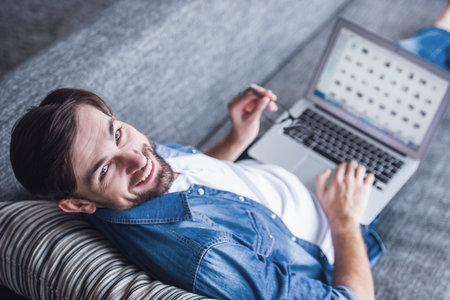 Handsome man is using a laptop, looking at camera and smiling while lying on couch at homeの写真素材
