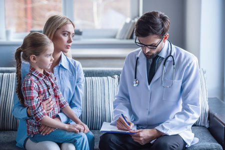 Beautiful young mother and her little daughter at the pediatrician. Doctor is making notesの写真素材