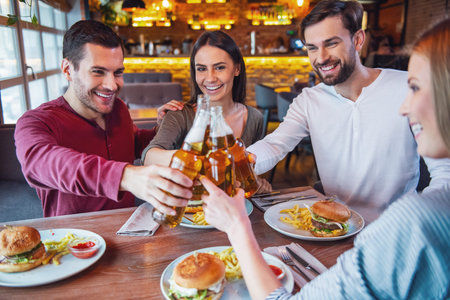 Happy friends are clinking bottles and smiling while spending time together in cafe