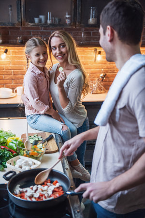 Cute little girl and her beautiful mom are  smiling while dad is cooking in kitchen at homeの写真素材