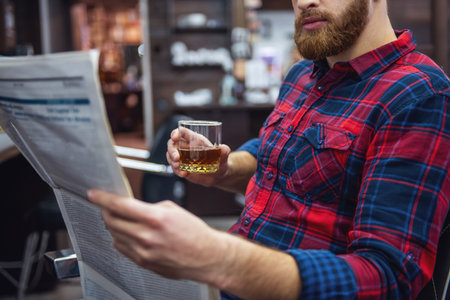 Handsome bearded man is reading a newspaper and holding a glass of beverage while sitting in chair at the barbershopの写真素材