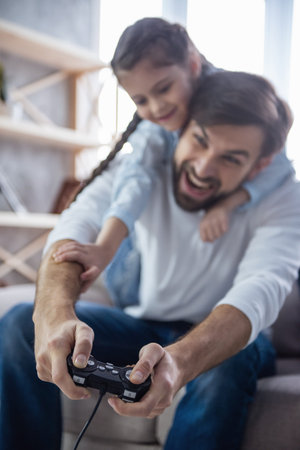 Cute little girl and her handsome father are playing game console and laughing while sitting on...