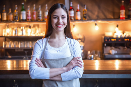Beautiful female barista is looking at camera and smiling while standing with folded arms near the bar counter in cafeの写真素材