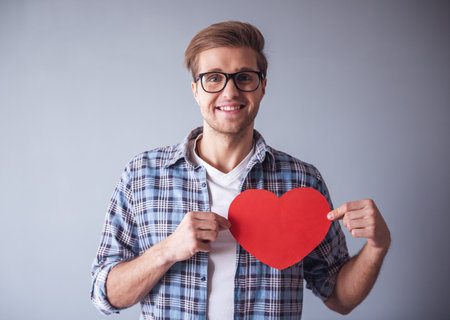 Handsome romantic guy is looking at camera and smiling while holding a red paper heart, on gray backgroundの写真素材