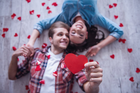 Top view of happy young couple looking at camera and smiling while lying on wooden floor. Guy is holding a red paper heartの写真素材