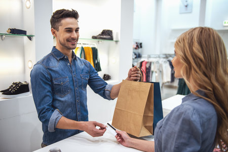 Handsome shop assistant is smiling while giving purchases and credit card to beautiful clientの写真素材