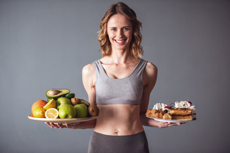 Beautiful young sportswoman is holding a tray with fruits and a plate with cakes, looking at camera and smiling, on gray backgroundの写真素材