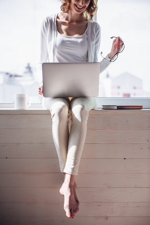 Beautiful woman is using a laptop and smiling while sitting on the window sill at homeの写真素材