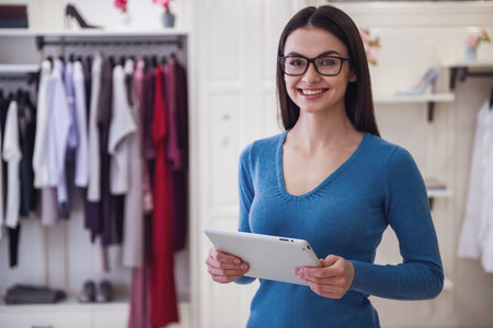 Beautiful female shop assistant in eyeglasses is using a digital tablet, looking at camera and smiling while standing in boutiqueの写真素材