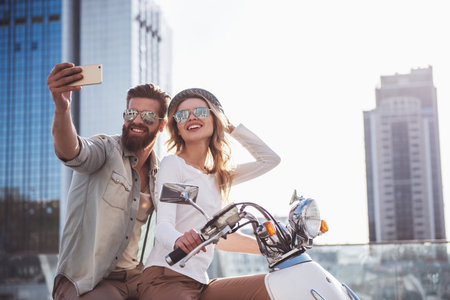 Beautiful young couple in sun glasses is doing selfie using a smart phone and smiling while sitting...