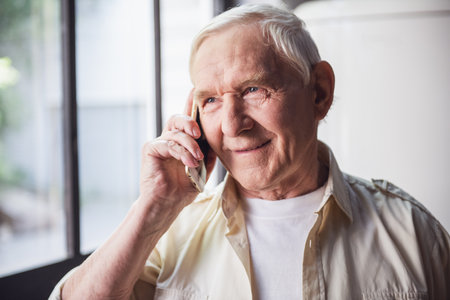 Handsome senior man is talking on the mobile phone and smiling while standing in kitchenの写真素材