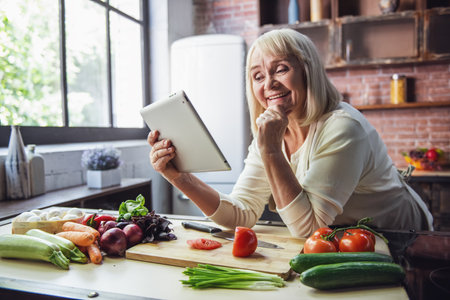 Beautiful senior woman is using a digital tablet, looking at camera and smiling while cooking in kitchenの写真素材