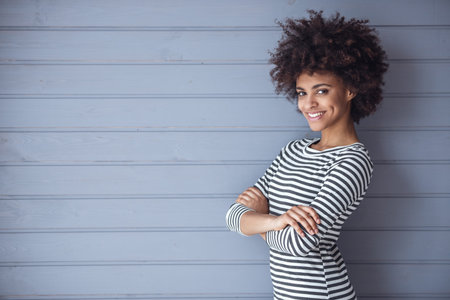 Stylish girl is looking at camera and smiling while standing with crossed arms on gray wall...