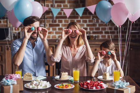 Family celebrating birthday in decorated kitchen. Mom, dad and son are holding macarons and smilingの写真素材