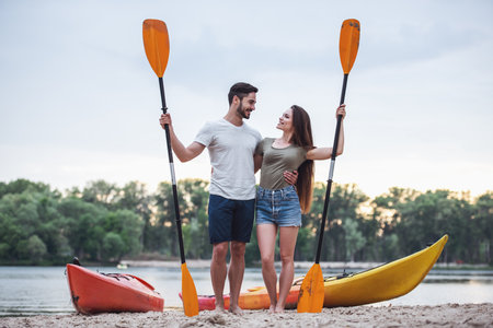 Happy young couple is holding paddles and smiling while standing near the kayaks on the beach after sailingの写真素材