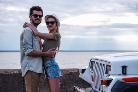 Beautiful couple is hugging, looking at camera and smiling while resting near the sea during their car tripの写真素材