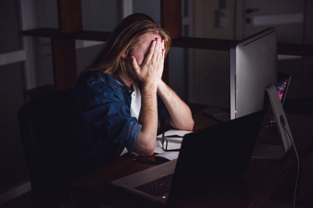 Handsome young businessman with shoulder-length blond hair is covering his face feeling tired working with a computer at nightの写真素材