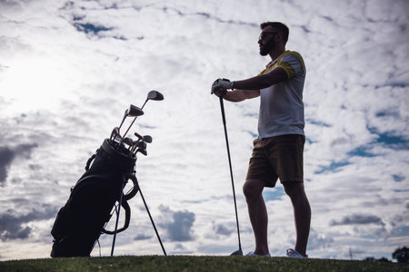 Full length portrait of handsome guy holding a golf club and looking away while standing on golf courseの写真素材