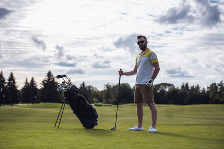 Full length portrait of handsome guy holding a golf club and looking at camera while standing on golf courseの写真素材
