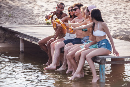 Happy friends are clinking bottles of soda water and smiling while sitting on pier at the seaの写真素材