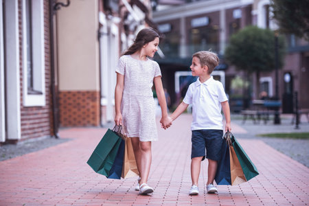 Cute little kids are carrying shopping bags, holding hands and smiling while walking down the streetの写真素材