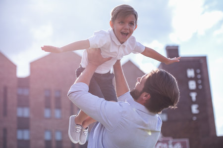 Handsome bearded dad is holding in arms his cute little son, who is flying and smiling while they are playing together outsideの写真素材