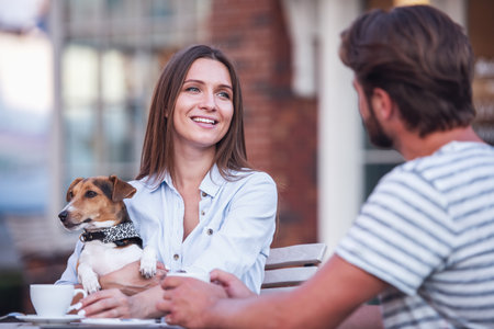 Beautiful woman is holding her cute dog, drinking coffee and smiling while talking with man in cafeの写真素材
