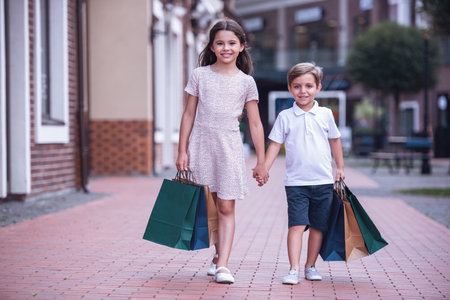 Cute little kids are carrying shopping bags, holding hands and smiling while walking down the streetの写真素材