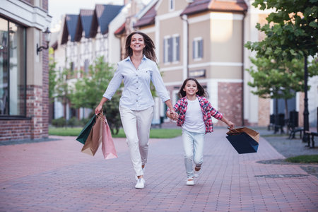 Beautiful mom and her cute little daughter are carrying shopping bags, holding hands and smiling while walking down the streetの写真素材