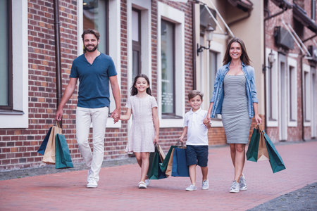 Beautiful parents and their cute little kids are carrying shopping bags, holding hands and smiling while walking down the streetの写真素材