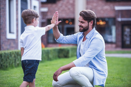 Handsome bearded dad is giving his cute little son a high five and smiling while they are playing together outsideの写真素材