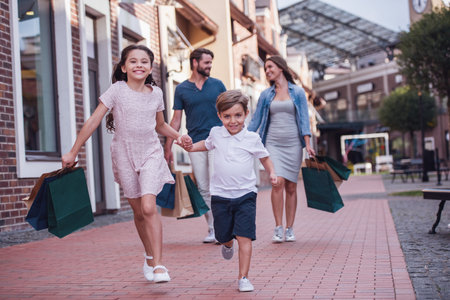 Beautiful parents and their cute little kids are carrying shopping bags, holding hands and smiling while walking down the streetの写真素材