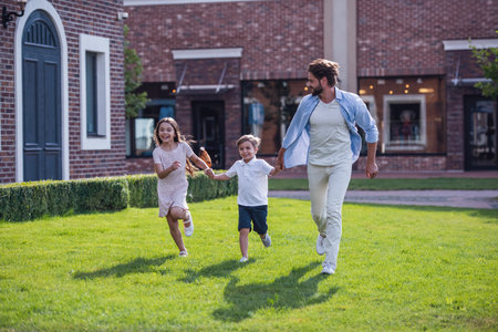 Cheerful kids and their handsome bearded father are holding hands, running and smiling while playing together outdoorsの写真素材