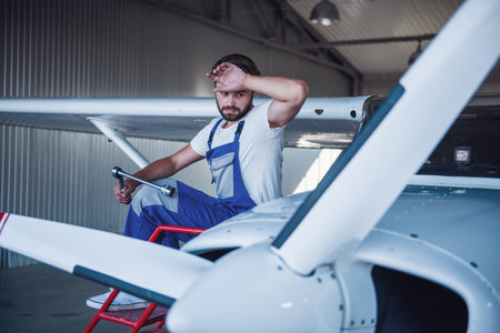 Handsome bearded mechanic in uniform is wiping his forehead while repairing an aircraft in hangarの写真素材