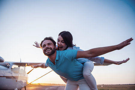 Beautiful romantic couple is looking away and smiling while standing on take-off ground near the aircraft, girl is piggybackの写真素材